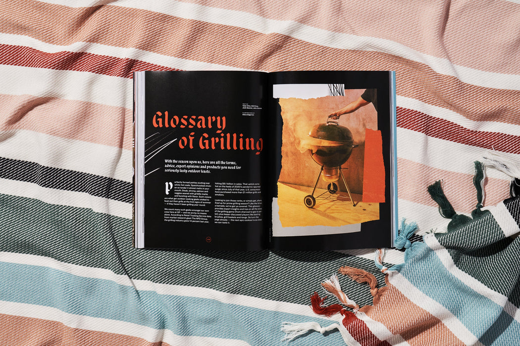 a copy of gear patrol magazine on a striped beach blanket open to the story glossary of grilling with a large visual of a charcoal grill