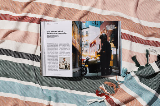 a copy of gear patrol magazine on a striped beach blanket open to the story Zen and the Art of Motorcycle Innovation with a large visual of a person near a motorcycle